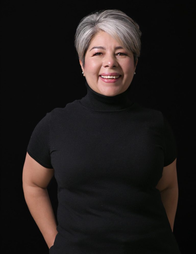 Mujer sonriente con cabello corto y canoso, vestida con camiseta negra. Fondo oscuro. Es Enid Alvarez Soberanis